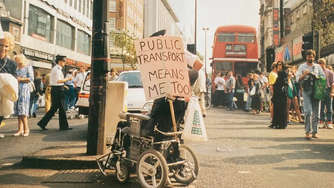 Image of disabled person in 1990s, protesting with plaque saying public transport means me too
