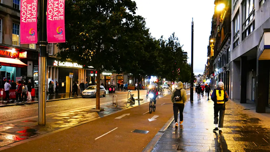 The western end of Sauchiehall Street at dusk: on the right people are walking and cycling by a People Make Glasgow banner and on the left a group of people are standing outside a bar