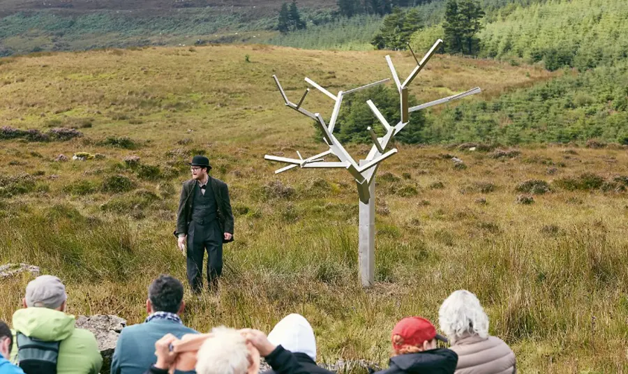 A man standing in a field in front of a tree sculpture. In the foreground are the back of the heads of an audience, watching the man.