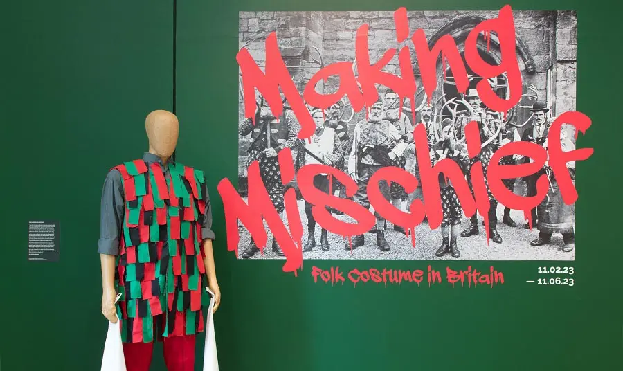 A mannequin wearing a bright green and black folk costume standing in front of a sign that reads 'Made in Mischief, Folk Costume in Britain'