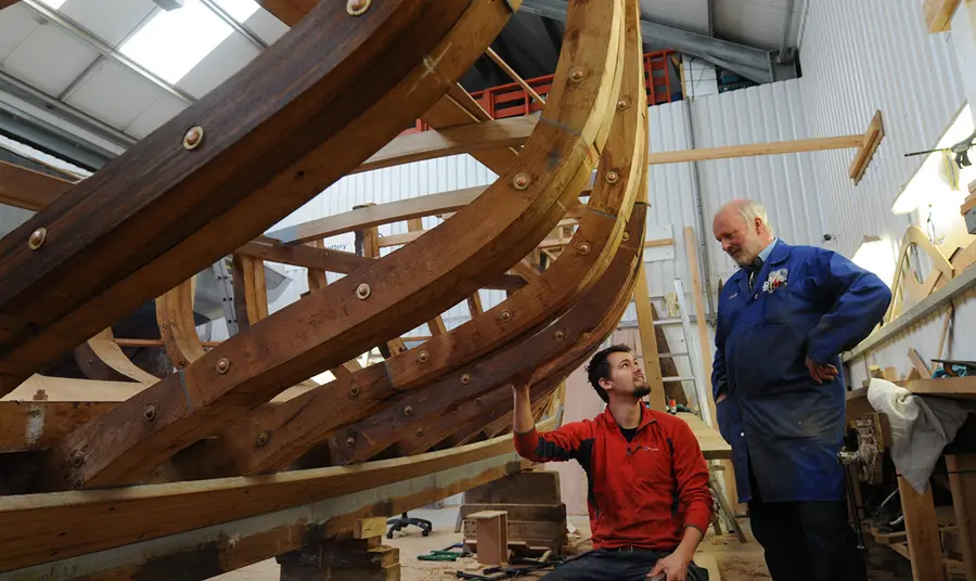 Two people stand next to the timber of a boat being built.