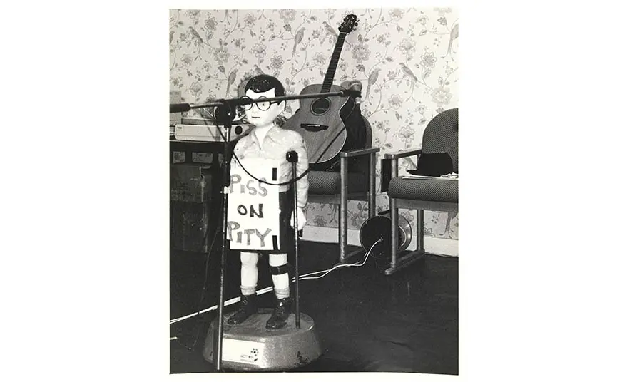 A black and white photo of a room set up for a concert, with a microphone and guitar visible. A plastic collection box in the shape of a disabled child is in the foreground, wearing a placard with the phrase 'piss on pity'
