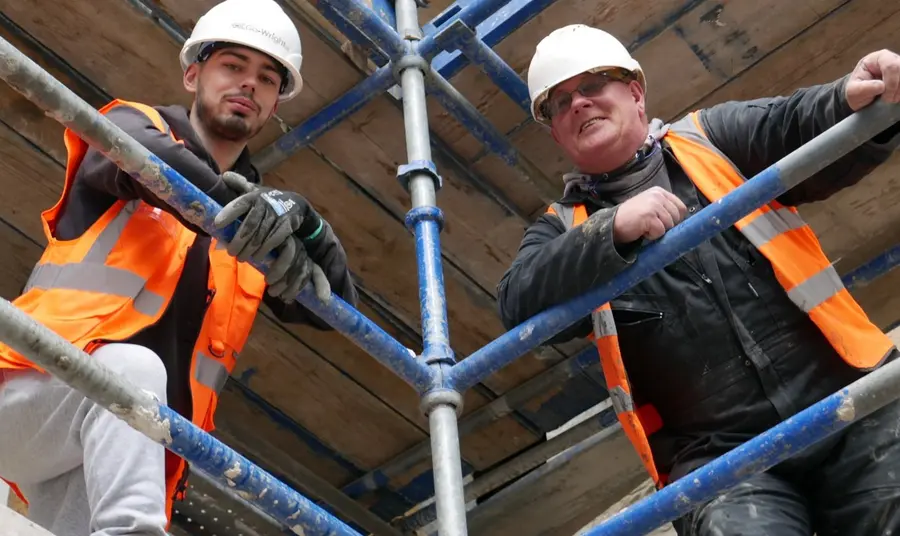 Two men wearing orange hi-viz vests and white safety helmets looking down towards the camera from scaffolding. On the left Jordan Docherty and on the right Stevie Pollock.