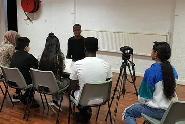 A group of young people sit at a table interviewing another young person, while another person films the interview