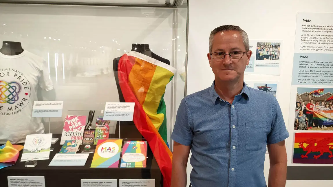 A person standing in front of a glass display of pride flags, t-shirts, leaflets and other memorabilia in a glass case