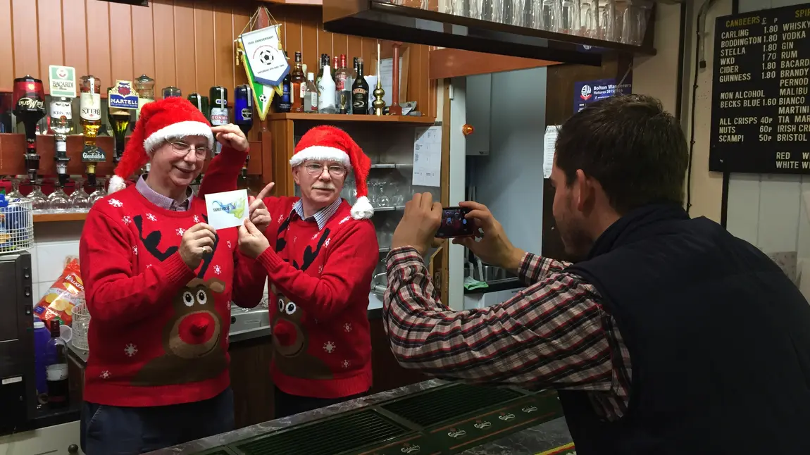 A person taking a photo of two people behind a bar wearing matching red Christmas hats and jumpers.