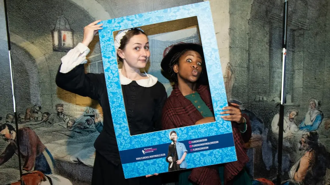 Two people in historic nursing outfits depicting Mary Seacole and Florence Nightingale, holding a frame to their faces