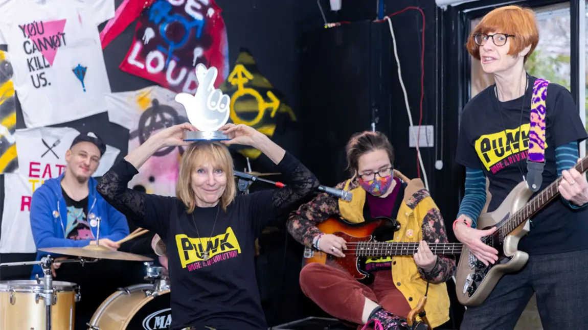 Four people play musical instruments including the drums and guitar in front of memorabilia from the Punk era