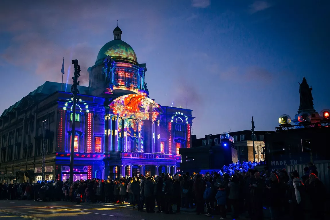 Colourful lights being projected onto a large historic building in a town square, in front of a large crowd